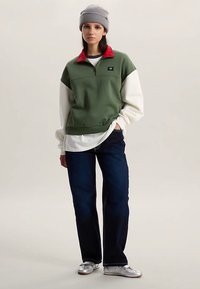 Grön sweatshirt med röd krage, vita ärmar och en liten varumärkestagg. Matchas med mörkblå jeans och silverfärgade skor. Mjuk textur, ledig passform.