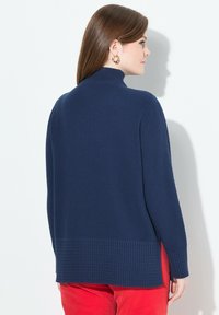 Frau in einem strukturierten, marineblauen Langarmpullover und roten Hosen, die von hinten blickt und sichtbare goldene Creolen trägt.