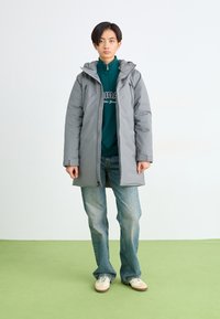 Grå isoleret parka med hætte, frontlynlås og to sidelommer, båret over en grøn lynlås-sweatshirt og blå jeans.