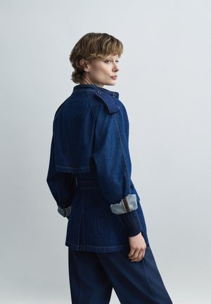 Άτομο που κοιτάζει πάνω από τον ώμο, φορώντας σκούρο μπλε τζιν μπουφάν με γυρισμένα μανίκια και σκούρο παντελόνι, μπροστά σε απλό ανοιχτόχρωμο φόντο.