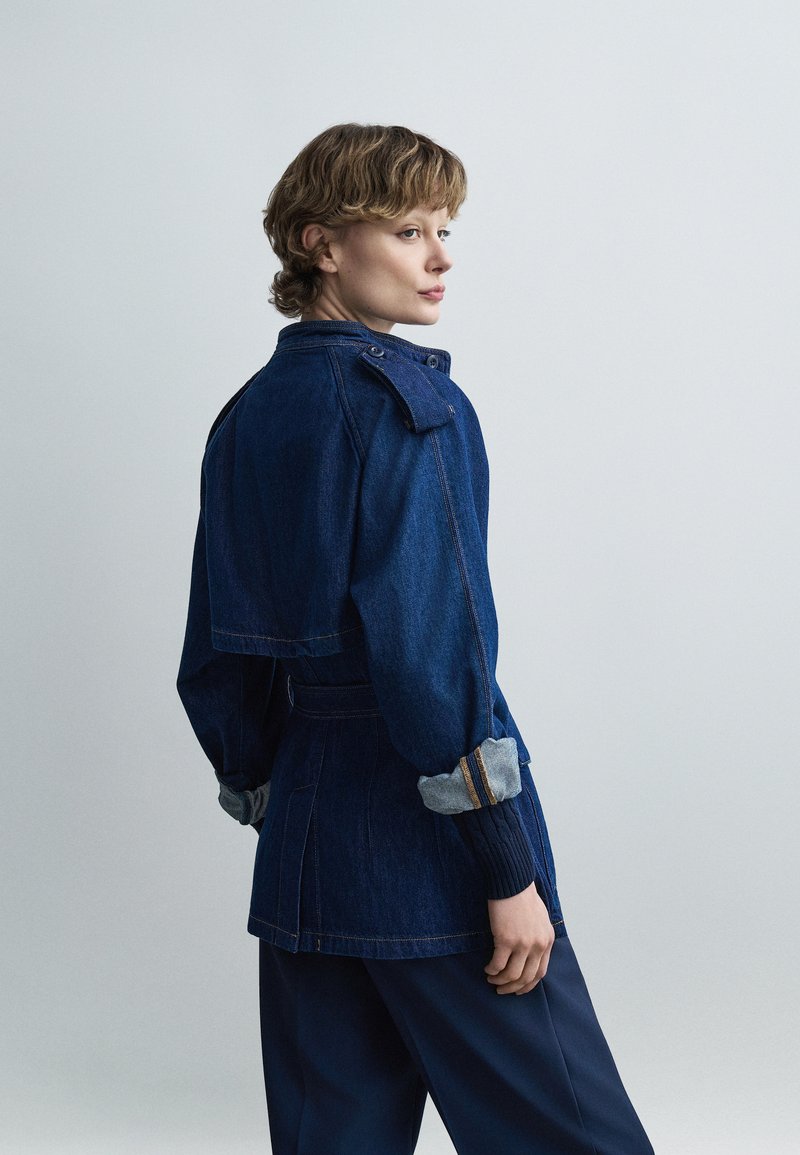 Άτομο που κοιτάζει πάνω από τον ώμο, φορώντας σκούρο μπλε τζιν μπουφάν με γυρισμένα μανίκια και σκούρο παντελόνι, μπροστά σε απλό ανοιχτόχρωμο φόντο.