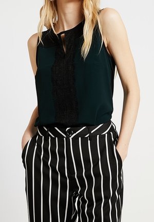 Blusa senza maniche verde scuro con dettagli in pizzo nero e scollatura a chiave, abbinata a pantaloni a vita alta a righe verticali bianchi e neri.