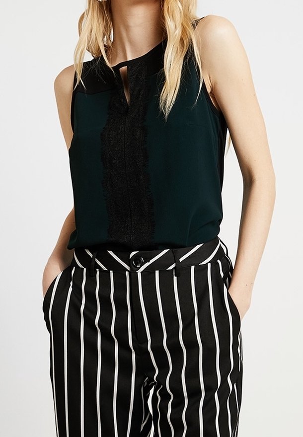 Ciemnozielona bezrękawnik z czarnym koronkowym detalem i dekoltem w kształcie klucza, w połączeniu z czarno-białymi spodniami o wysokim stanie w pionowe paski.