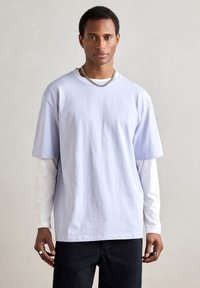 Világoslila, túlméretezett rövid ujjú póló, fehér hosszú ujjú ing fölött rétegezve. A modell ezüst lánc nyakláncot visel.