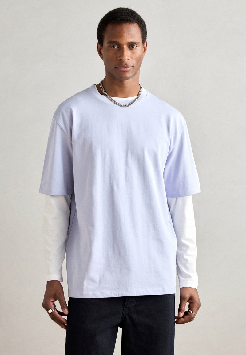 Világoslila, túlméretezett rövid ujjú póló, fehér hosszú ujjú ing fölött rétegezve. A modell ezüst lánc nyakláncot visel.