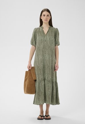 Nő zöld pöttyös midi ruhában, nagy barna szarvasbőr táskát tartva, fekete lapos szandált viselve, egyszínű fehér háttér előtt áll.