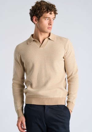 Joven con cabello rizado que lleva un suéter tipo polo de manga larga beige con rayas y pantalones oscuros, mirando hacia su derecha con las manos en los bolsillos.