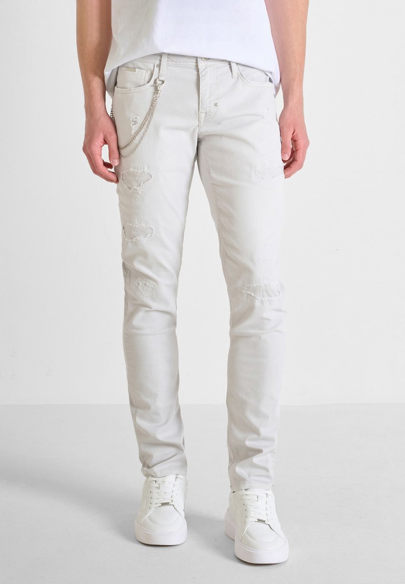 Jeans blancos de corte slim con parches desgastados, diseño de cinco bolsillos y un detalle de cadena en el lateral. Combinados con zapatillas blancas. Textura suave.