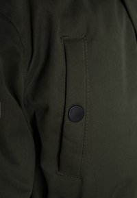 Tissu vert foncé avec une texture lisse, comportant une poche à rabat et un bouton noir étiqueté "Tom Tailor". Couture visible.