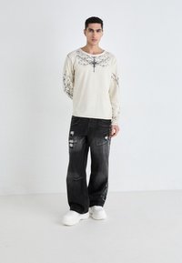 Camisa de manga larga beige con estampado floral negro, combinada con jeans negros de corte holgado con detalles desgastados, y zapatillas blancas gruesas.