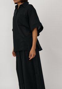 Kobieta w luźnej czarnej koszuli z krótkim rękawem i kołnierzykiem oraz dopasowanych szerokich spodniach, stojąca na jasnym, jednolitym tle.