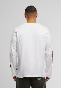 Long-sleeve white t-shirt with a crew neck and graphic prints on the sleeves; paired with dark jeans featuring stitched details.