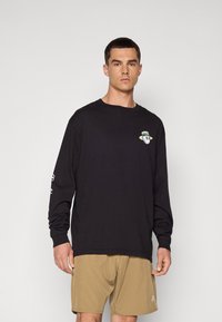 Black long-sleeve T-shirt with a round neckline, featuring a graphic of a character wearing a hat. Paired with tan shorts.
