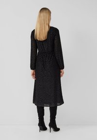 Kobieta z długimi blond włosami stoi w czarnej, midi sukience z długim rękawem o teksturowanym wzorze oraz czarnych wysokich obcasach na jednolitym tle.