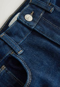 Nahaufnahme einer dunkelblauen Denim-Jeans mit Gürtelschlaufen, Kanten der Tasche und einem metallischen Knopf mit der Aufschrift "Premium Collection".