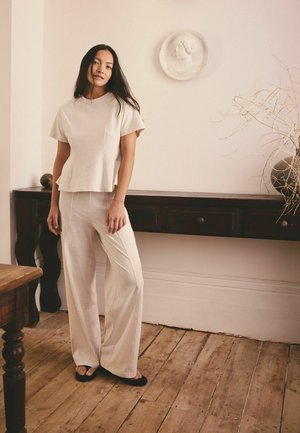 Frau steht drinnen und trägt beige, weit geschnittene Hosen und ein passendes kurzärmeliges Oberteil, im Hintergrund dunkle Holzmöbel und Wanddekoration.