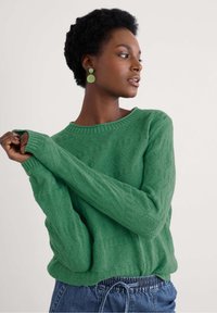 Zaļš adīts džemperis ar teksturētu dimanta rakstu, apaļu kakla izgriezumu un garām piedurknēm, pārī ar denima biksēm.