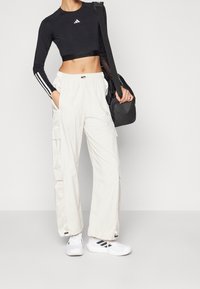 Černý crop top s dlouhým rukávem, bílými pruhy a logem, v kombinaci se světle béžovými cargo kalhotami s kapsami, obutý s bílými sportovními botami.