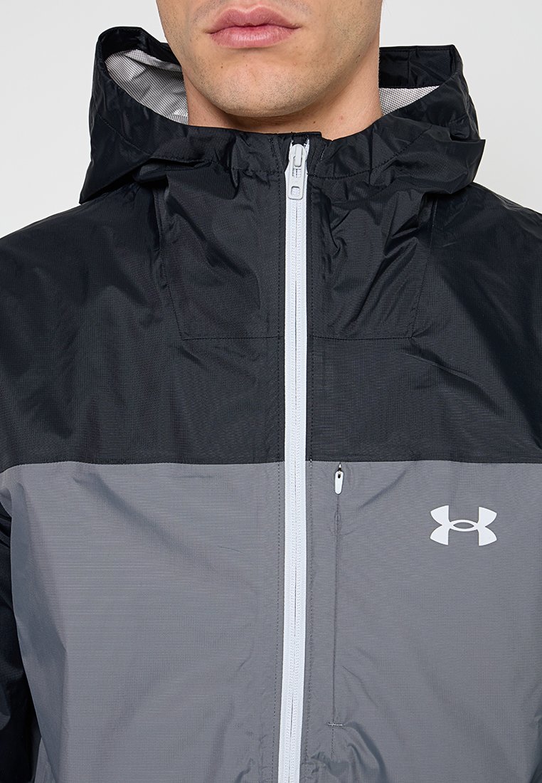 Chaqueta ligera en negro y gris con capucha, que cuenta con un cierre frontal, forro de malla y un logo blanco en la parte inferior derecha.