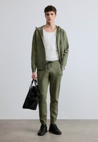 Chándal deportivo verde oliva con textura acanalada, sudadera con capucha y cremallera frontal, pantalones con cordón ajustable y zapatillas negras. Bolsa negra en la mano.