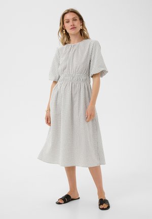 Femme portant une robe midi à carreaux blancs et noirs et des sandales noires, debout devant un fond blanc uni.