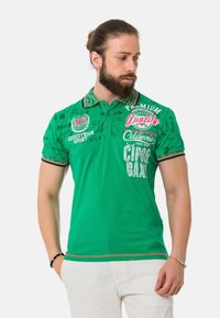 Grünes Polo-Shirt aus Baumwolle mit Kragen, kurzen Ärmeln und grafischen Drucken in Weiß und Rot. Auffällige Akzente sind die gestreiften Ränder.