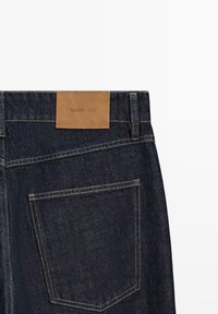 Dark blue denim jeans back pocket with contrasting yellow stitching and a tan leather patch labeled "Massimo Dutti" at the waistband.