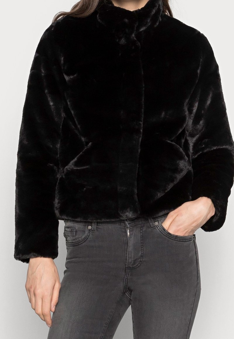 Femme portant une veste en fausse fourrure noire à manches longues et un jean gris foncé, une main dans la poche et l'autre détendue à côté.