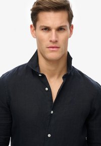 Donkerblauw linnen shirt met een kraag, voorzien van een knoopsluiting en een subtiel geborduurd logo op de linkerborst. Soepele textuur.