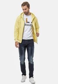 Geel zip-up hoodie over een wit T-shirt met een kleurrijke opdruk en zwarte tekst, gecombineerd met een donkere spijkerbroek en witte sneakers.