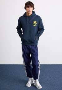Mörkblå hoodie med gul grafik, parat med mörkblå träningsbyxor med vita ränder. Klädseln inkluderar grå sneakers och en axelrem.
