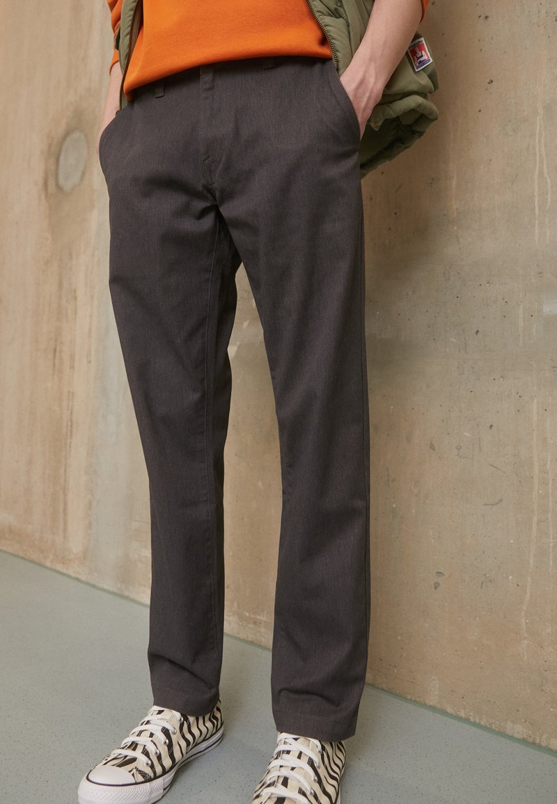 Pantalons ajustés gris avec une texture lisse, comprenant des poches avant et un design coupe droite, associés à des sneakers rayées blanches et noires.