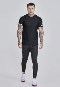 Fekete sportpóló rövid ujjakkal, sima textúrával; slim-fit fekete nadrággal és fehér sneakers-szel, melyen lila részletek találhatók.