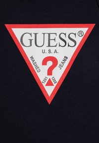 T-shirt preta com um grande logótipo triangular vermelho e branco. O texto inclui "GUESS", "U.S.A.", "WASHED", "JEANS" e um ponto de interrogação central.