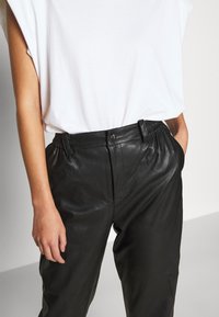 Kvinna som bär svarta högmidjade läderbyxor med elastiska sidor och en knapp, kombinerat med en lös vit ärmlös topp, händerna i fickorna.