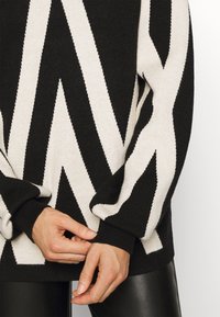 Schwarzer und cremefarbener Pullover mit Zickzackmuster, aus strukturiertem Stoff gefertigt. Rippbündchen und Saum sorgen für eine enganliegende Passform. Nahaufnahme von Händen, die den Ärmel anpassen.