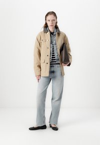 Béžová oversize bunda s šedým límcem, modrá denimová bunda pod ní, pruhovaná košile, světle modré džíny a černé ploché boty. Drží hnědou tašku.