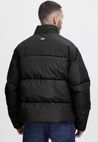 Black puffer jacket with a high collar, quilted design, and stitched panels. Features a logo patch on the back. Textured, insulated material.