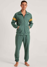 Groene trainingsoutfit van zachte stof, bestaande uit een ritsjack met een kraag en gele schouderstrepen, gecombineerd met bijpassende broek.