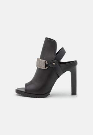 Black open-toe heeled shoe with a smooth leather upper, elastic band detail, and a textured rubber sole. Slim stiletto heel design.