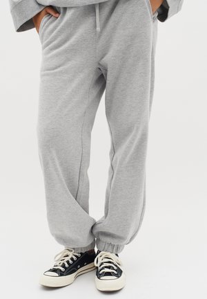 Pantalons de survêtement gris en matériau doux, avec des poignets élastiques et des poches latérales, associés à des baskets noires avec des semelles et des lacets blancs.