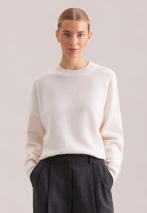 Frau mit zurückgestrichenem Haar, die einen weißen Strickpullover und dunkelgraue, hoch taillierte Hose trägt, mit den Händen in den Taschen vor einem schlichten Hintergrund.