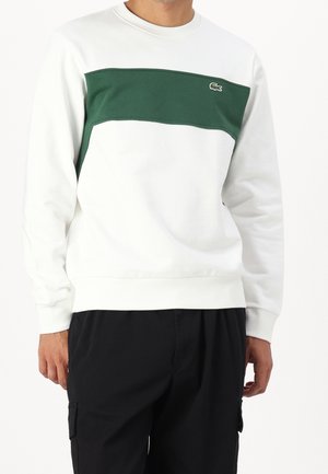 Mies, joka pukeutunut valkoiseen svetariin, jossa on tummanvihreä vaakasuora paneeli ja pieni krokotiililogolla, yhdistetty mustiin housuihin.