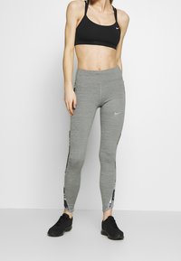 Leggings grigi con accenti neri sui lati e logo Nike, abbinati a un reggiseno sportivo nero. Il materiale sembra elastico e comodo.