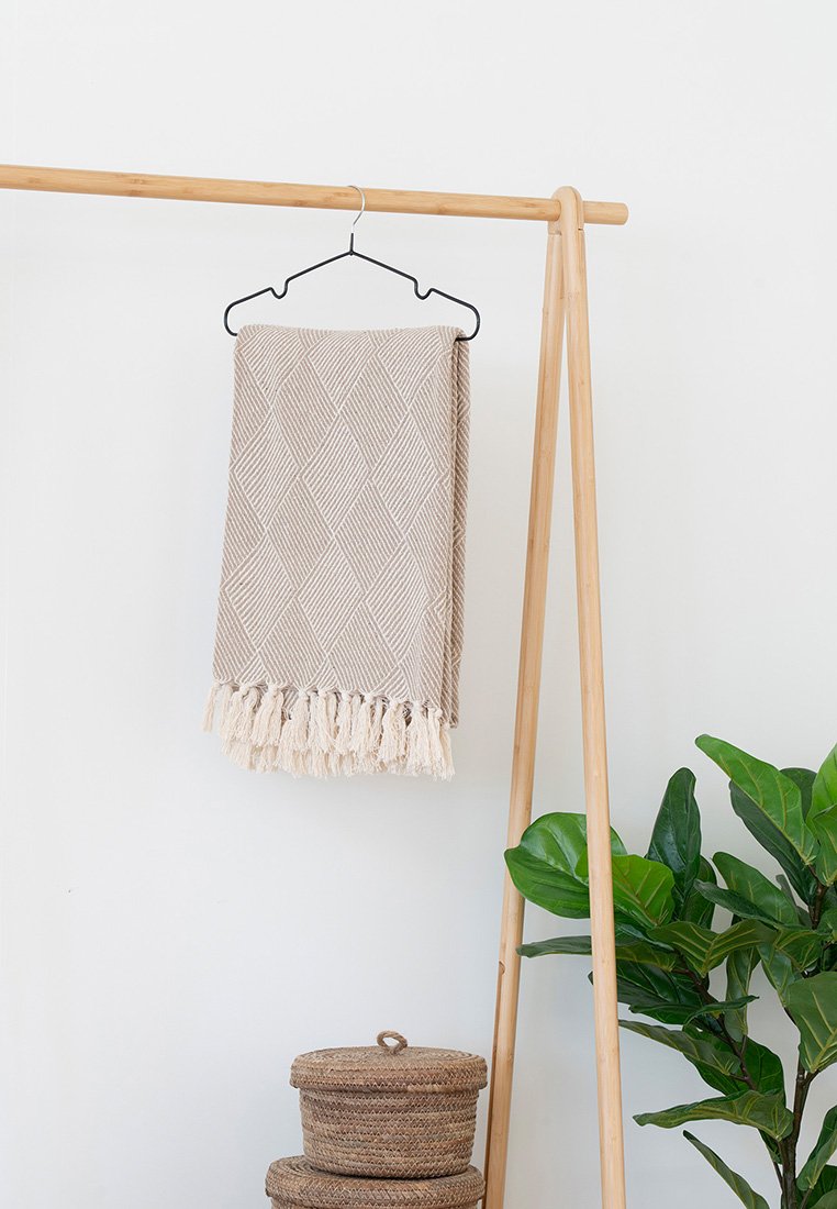 Coperta intrecciata beige con un motivo a rombi, bordi frangiati, appesa a un gancio nero su un supporto di legno, accanto a una pianta verde.