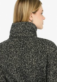 Cappotto in misto lana con colletto alto, caratterizzato da un motivo a spina di pesce nei colori nero e crema. Superficie texturizzata con vestibilità rilassata.
