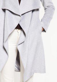Cappotto in lana grigio chiaro con frontale drappeggiato e collo sciallato, dotato di maniche lunghe e tasche laterali, indossato sopra un top marrone e pantaloni bianchi.