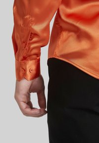 Chemise orange en satin à manches longues avec poignets boutonnés et texture lisse, présentée sur un fond gris, montrant partiellement une main.