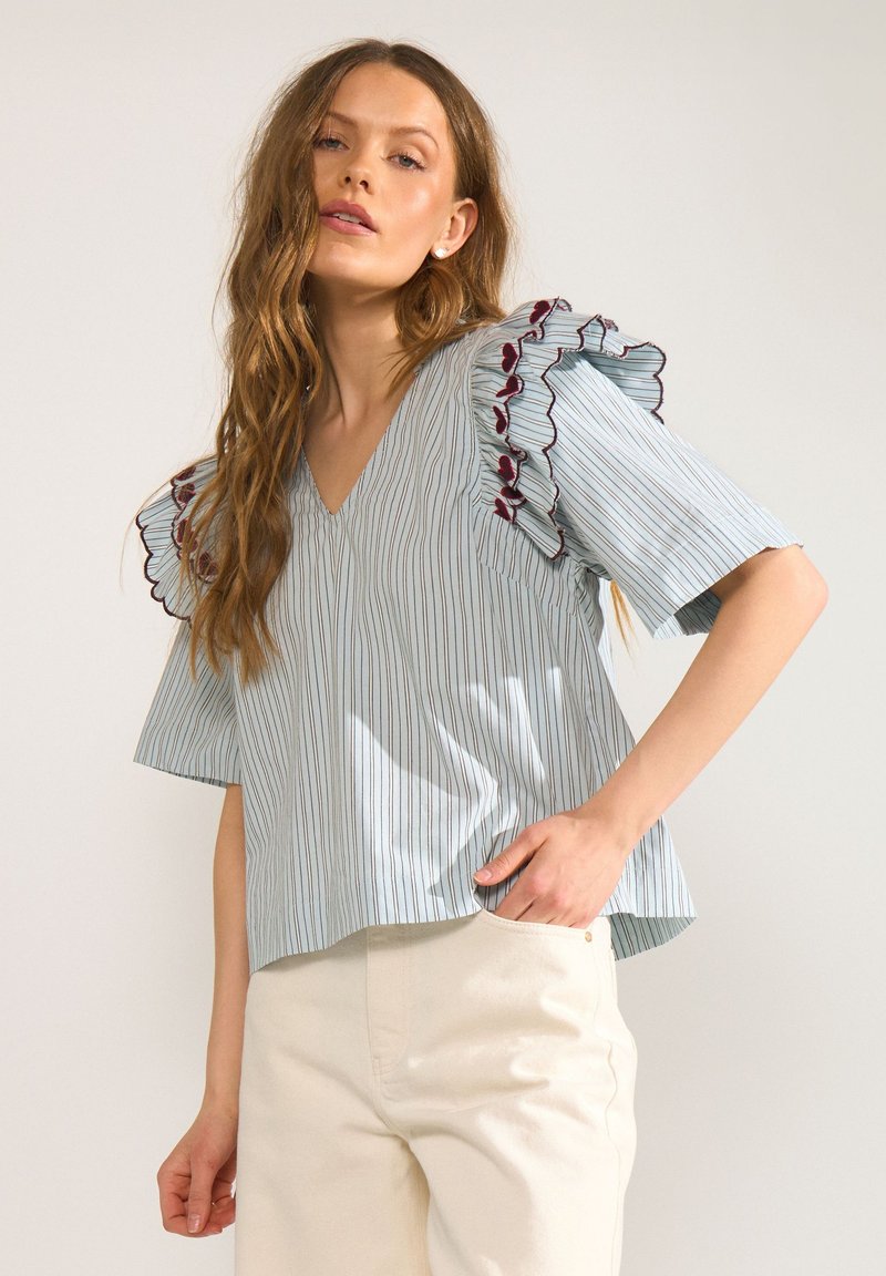 Femme aux longs cheveux ondulés, portant un chemisier rayé bleu clair avec des manches à volants et un pantalon couleur crème, posant avec une main dans la poche.