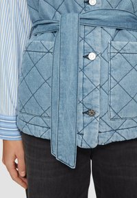 Gepunktete Jeansweste in Hellblau mit einem Bindegürtel, silbernen Druckknöpfen und zwei Fronttaschen. Die darunter liegende Schicht hat ein blau-weiß gestreiftes Hemd.